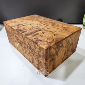 Könnte beinhalten: Eine rechteckige Holzkiste mit Scharnierdeckel. Die Kiste besteht aus hellbraunem Holz mit einem einzigartigen, wirbelnden Maserungsmuster. Sie hat einen kleinen, goldfarbenen Verschluss auf der Vorderseite. Die Kiste dient wahrscheinlich zur Aufbewahrung oder Präsentation.