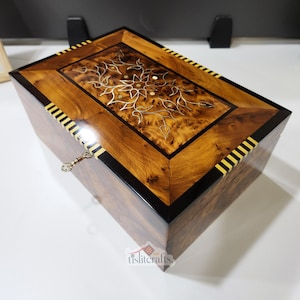 Op de afbeelding: Een rechthoekige houten kist met een scharnierend deksel. De kist heeft een ingelegd ontwerp op het deksel en een sleutelgat met een kleine sleutel. Het hout heeft een warme tint met donkere en lichte accenten. Ongeveer 25 cm lang.