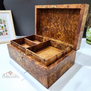 Op de afbeelding: Open houten sieradenkist met een patroon aan de buitenkant. De doos heeft een binnenvak met compartimenten. Een ingelijste foto van planten en een kleine potplant staan op de achtergrond.