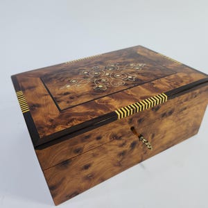 Op de afbeelding: Een rechthoekige houten kist met een sleutelgat en een decoratief ingelegd ontwerp op het deksel. De kist heeft een rijke, warme houtnerf met donkerbruine randen en gele en gouden accenten. De kist is ongeveer 30 cm lang.