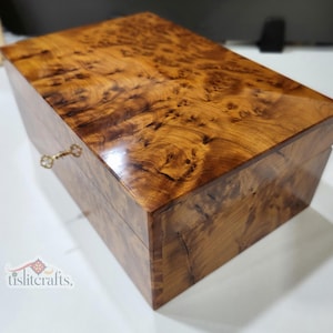 Può includere: Una scatola rettangolare in legno con finitura lucida. La scatola è realizzata in legno marrone chiaro con un motivo a venature unico e vorticoso. Una piccola serratura e una chiave dorate e ornate sono visibili sulla parte anteriore.