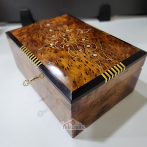 Op de afbeelding: Een houten sieradendoos met een sleutelslot. De doos is gemaakt van donker hout met een wortelnotenpatroon en heeft een decoratieve inleg op het deksel. Het deksel heeft een zwarte rand met gouden strepen.