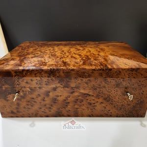 Puede incluir: Una caja rectangular de madera con un rico acabado de veta de madera de raíz. La caja tiene un brillo brillante y está adornada con pequeños herrajes dorados ornamentados. La paleta de colores general es cálida, con tonos marrones y ámbar.