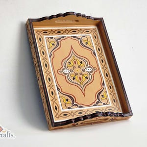 Op de afbeelding: Rechthoekig houten dienblad met een decoratief, symmetrisch ontwerp. De lade heeft een lichtbruine achtergrond met een centraal patroon in geel, wit en bruin. De randen zijn donkerbruin met een geschulpte rand.