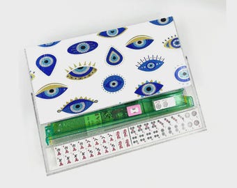 May include: A clear acrylic Mahjong set with a white lid featuring a blue and gold evil eye pattern. The set includes green dice, and white and red tiles. The set is displayed on a white background.