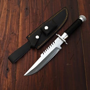 Può includere: Un coltello da sopravvivenza con manico nero e lama seghettata, accanto a un fodero in pelle nera con passante per cintura. Il coltello ha una guardia argentata e un tappo terminale argentato strutturato. Il fodero ha un bordo cucito.