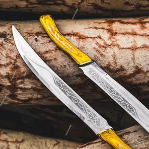 May include: Two silver blades with ornate etched designs and yellow wooden handles. The blades are angled towards the viewer, with the handles at the top of the frame. The background is a textured, brown surface.