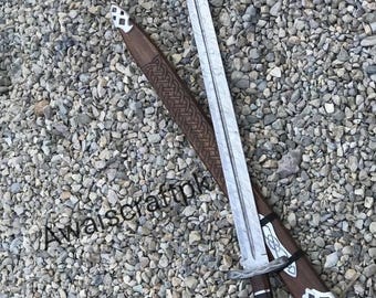May include: A silver-colored sword with a pointed tip and a brown handle rests on a brown sheath with a decorative pattern. The sword has a detailed hilt and a white and black design. The background is a bed of small gray stones.