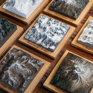 May include: A selection of 3D topographical models of famous mountains, including Mount Everest and Kilimanjaro, presented on wooden bases. Each model is labelled with the mountain's name and elevation in metres. The models feature detailed textures and realistic colouring.