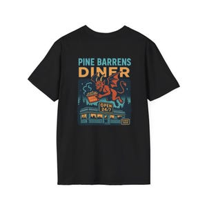 Może przedstawiać: Czarny t-shirt z grafiką czerwonego diabła trzymającego frytki przed restauracją. Tekst na koszulce brzmi "PINE BARRENS DINER" i "OPEN 24/7". Grafika restauracji zawiera las i słowa "Since 1909".