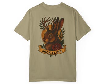 Könnte beinhalten: Khaki-farbenes T-Shirt mit einer detaillierten Illustration eines Jackalopen. Das Design zeigt einen braunen Jackalopenkopf mit Geweih, eine rote Rose und das Wort "JACKALOPE" auf einem Banner.