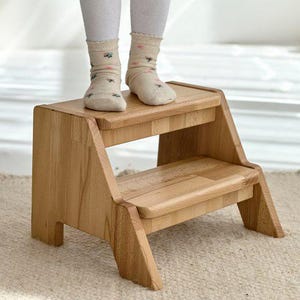 Pode incluir: Um banco de madeira de dois degraus com acabamento natural. O banco tem um degrau superior retangular e um degrau inferior, com suportes laterais angulados. A imagem mostra as pernas e os pés de uma pessoa em pé no banco.