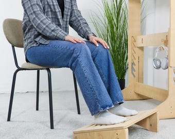 May include: A light-colored wooden footrest with a slatted top is positioned in front of a person wearing blue jeans and white socks. The footrest is part of a larger wooden desk setup. A person is sitting on a chair.