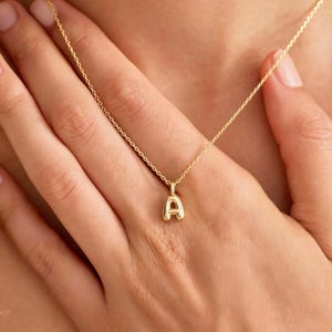Puede incluir: Un collar dorado con un colgante con la forma de la letra "A". El colgante tiene un diseño redondeado en forma de burbuja. El collar está suspendido de una fina cadena. La imagen muestra el collar sobre la piel de una persona.