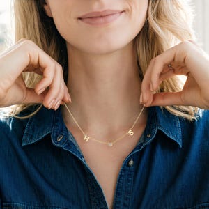 Puede incluir: Un delicado collar de oro con las letras "H" y "S" y un pequeño colgante de diamante. El collar se muestra sobre una camisa vaquera.