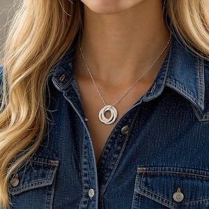 Puede incluir: Collar plateado con un colgante de tres anillos entrelazados. El collar se luce sobre una camisa vaquera. Los anillos tienen un acabado pulido, ofreciendo un diseño sencillo pero elegante. La cadena es fina, realzando la apariencia del colgante.