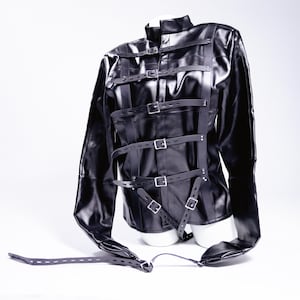 Puede incluir: Chaqueta de cuero negro con múltiples correas y hebillas plateadas. La chaqueta tiene mangas largas y un cuello alto. Las correas están diseñadas para asegurar al usuario. La chaqueta se exhibe en un maniquí blanco.