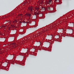 Puede incluir: Dos tiras de encaje rojo con acabado brillante. El encaje tiene un borde festoneado con calados en forma de diamante. El material parece ser tela, sobre un fondo blanco.
