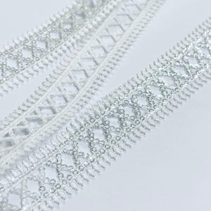 Op de afbeelding: Close-up van twee witte kanten linten met een zilveren metallic draad geweven in een geometrisch patroon. De linten liggen op een witte achtergrond. Het kant heeft een delicaat, decoratief ontwerp, geschikt voor knutselen en versieringen.