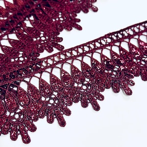 Può includere: Un bordo in pizzo bordeaux intenso caratterizzato da un intricato bordo smerlato e da un motivo ripetitivo a goccia. Il pizzo è impreziosito da piccole paillettes rosso scuro abbinate che offrono un sottile luccichio su tutta la superficie.