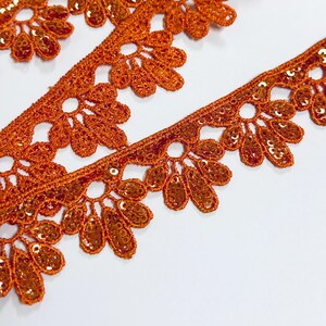 May include: A length of burnt orange lace trim featuring a repeating scalloped floral pattern. The intricate embroidery is embellished with small, matching orange sequins that catch the light, creating a textured and decorative border edge.