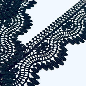 Può includere: Primo piano di un bordo in pizzo nero con un bordo smerlato e un motivo floreale. Il pizzo è realizzato in un materiale morbido e strutturato ed è posto su uno sfondo bianco. Il bordo è ideale per progetti di cucito e artigianato.
