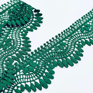 Könnte beinhalten: Nahaufnahme einer Rolle smaragdgrüner Spitzenborte. Die Borte weist ein detailliertes, offenes Design mit gewellten Kanten und blattartigen Mustern auf. Die Spitze wird vor einem weißen Hintergrund präsentiert, der ihre komplizierte Textur und lebendige Farbe hervorhebt.