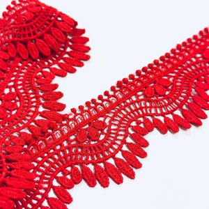 Può includere: Un rotolo di passamaneria in pizzo rosso vivo. Il pizzo presenta un design traforato dettagliato con un bordo smerlato ed elementi a forma di foglia. La passamaneria è esposta su uno sfondo bianco, evidenziando il suo motivo intricato.
