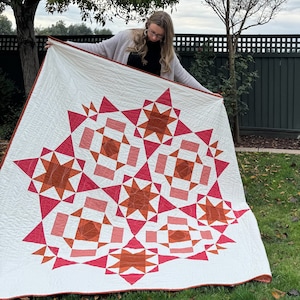 Puede incluir: Una colcha blanca con un patrón geométrico de estrellas y cuadrados rojos, naranjas y rosas. La colcha está siendo sostenida por una persona en un jardín.