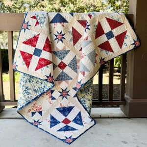 May include: A red, white, and blue patchwork quilt with a floral pattern. The quilt is folded and draped over a wooden railing.