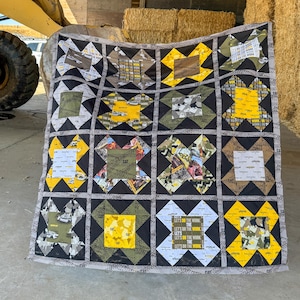 May include: A patchwork quilt with a repeating pattern of squares and diamonds. The quilt features black, gray, yellow, and camouflage fabric squares. Some squares have the text "LET'S DO THE WORK". The quilt is displayed in a barn.