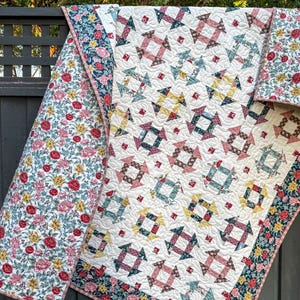 Puede incluir: Un edredón hecho a mano con un fondo color crema y un patrón geométrico en tonos rojos, azules y amarillos. El borde del edredón presenta un estampado floral con flores rojas, rosas y amarillas sobre un fondo azul marino. El edredón se exhibe al aire libre.