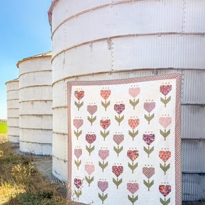 Può includere: Una trapunta bianca con un motivo floreale di fiori a forma di cuore nei toni del rosa e del bordeaux. La trapunta ha un bordo a scacchi bordeaux e bianco, appesa davanti a silos bianchi.