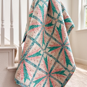 May include: A quilted blanket featuring a repeating starburst pattern in shades of teal, soft pink, and white. The fabric displays intricate geometric and floral motifs throughout the design, with visible stitching lines creating a textured surface.