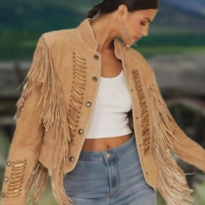 May include: A tan suede jacket with fringe detailing on the sleeves and back. The jacket features a round collar, button closures, and decorative stitching. The sleeves have fringe and metal accents. The jacket is paired with a white top and blue jeans.