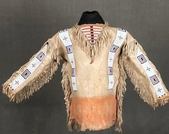 Puede incluir: Una camisa de manga larga de color beige con intrincados abalorios y flecos. La camisa presenta patrones geométricos en blanco, rojo y azul. El cuello es negro y la parte inferior es de un tono naranja más oscuro. Las mangas y los hombros están adornados con rayas de cuentas blancas.
