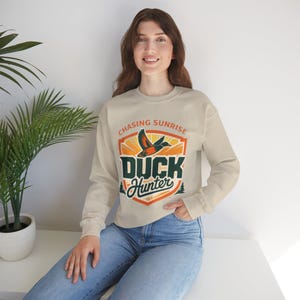 May include: A beige sweatshirt featuring the text "Chasing Sunrise" and "Duck Hunter" within a shield design. The graphic includes a flying duck, a rising sun, and pine trees. The sweatshirt has long sleeves.