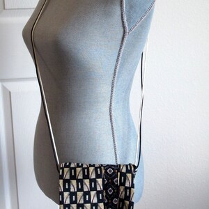 May include: A small, rectangular crossbody bag with a black, white, and gold geometric pattern. The bag is suspended from a thin, black and white strap. The bag is displayed on a mannequin.