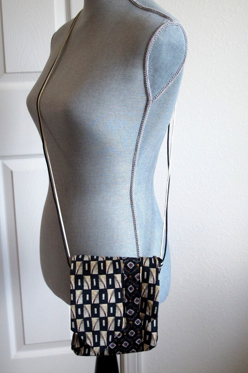 May include: A small, rectangular crossbody bag with a black, white, and gold geometric pattern. The bag is suspended from a thin, black and white strap. The bag is displayed on a mannequin.
