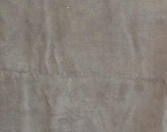May include: A close-up shot of a piece of fabric in a muted gray-green color. The fabric has a soft, suede-like texture with subtle variations in tone. The surface shows slight creases and folds, suggesting it is a pliable material.