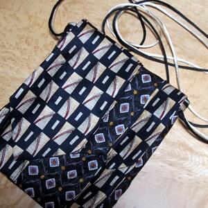 May include: A handmade crossbody bag crafted from repurposed neckties. The bag features a geometric pattern in navy, beige, and burgundy, with a contrasting floral design. It has a black and white cord strap.