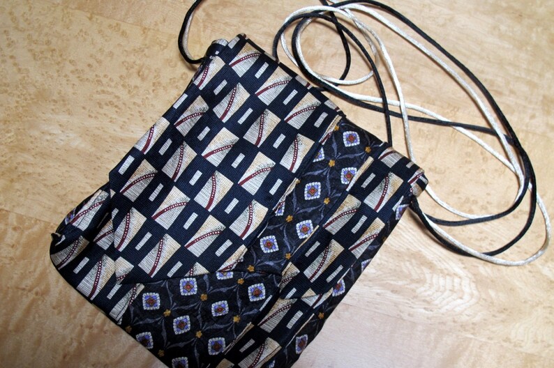 May include: A handmade crossbody bag crafted from repurposed neckties. The bag features a geometric pattern in navy, beige, and burgundy, with a contrasting floral design. It has a black and white cord strap.