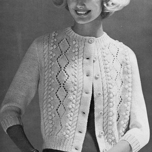 Puede incluir: Rebeca color crema con botones y patrones de punto intrincados. El suéter presenta diseños de diamantes y trenzas, con pequeñas bolitas que agregan textura. Las mangas son de tres cuartos, usado por una persona.