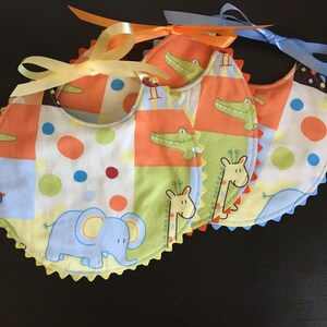 May include: Four baby bibs with colorful patchwork patterns. Each bib features a different animal: an elephant, a crocodile, and a giraffe. The bibs are trimmed with a scalloped edge and tied with a ribbon.