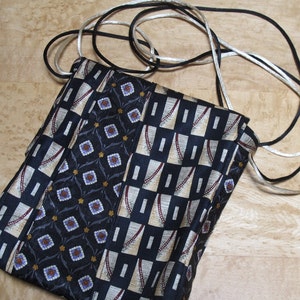 May include: A handmade crossbody bag crafted from repurposed neckties. The bag features a patchwork design with various patterned fabrics in navy, beige, and black. It has a long strap made of black and silver cord.