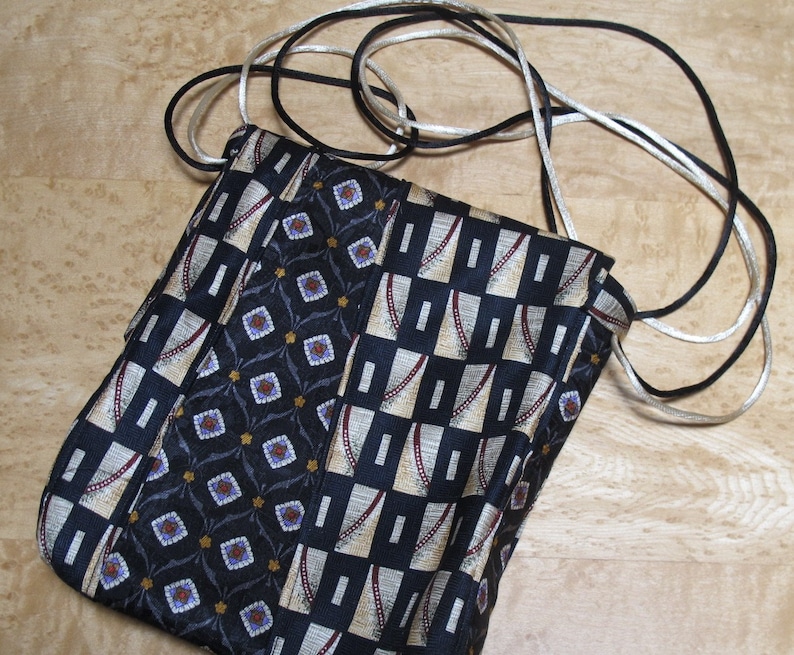 May include: A handmade crossbody bag crafted from repurposed neckties. The bag features a patchwork design with various patterned fabrics in navy, beige, and black. It has a long strap made of black and silver cord.