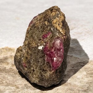 Può includere: Un esemplare minerale grezzo e non lucidato caratterizzato da una matrice scura, ruvida e grigio-marrone, con una prominente inclusione cristallina incastonata di rubino rosso magenta intenso. La pietra ha una forma irregolare e frastagliata.
