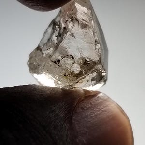 Puede incluir: Una gema transparente y en bruto, sostenida entre dos dedos. La gema tiene una estructura cristalina con inclusiones visibles y una forma ligeramente irregular. La luz atrapa las facetas, creando un efecto brillante. La apariencia general sugiere un diamante natural en bruto.