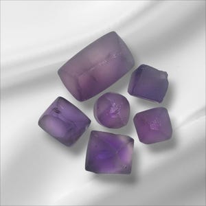 May include: A collection of translucent, purple crystals displayed on a white background. The crystals are various shapes, including cubes and rounded forms, with a soft, diffused light. The image evokes a sense of natural beauty and geological interest.