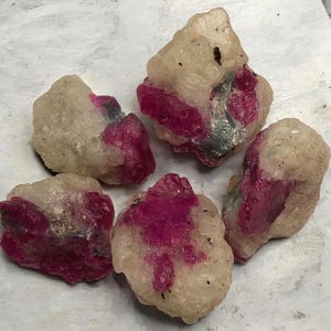 May include: Five raw, unpolished mineral specimens featuring a combination of translucent, off-white crystalline matrix and vibrant, deep magenta-colored ruby inclusions. Each stone has an irregular, jagged shape with a rough, natural texture.